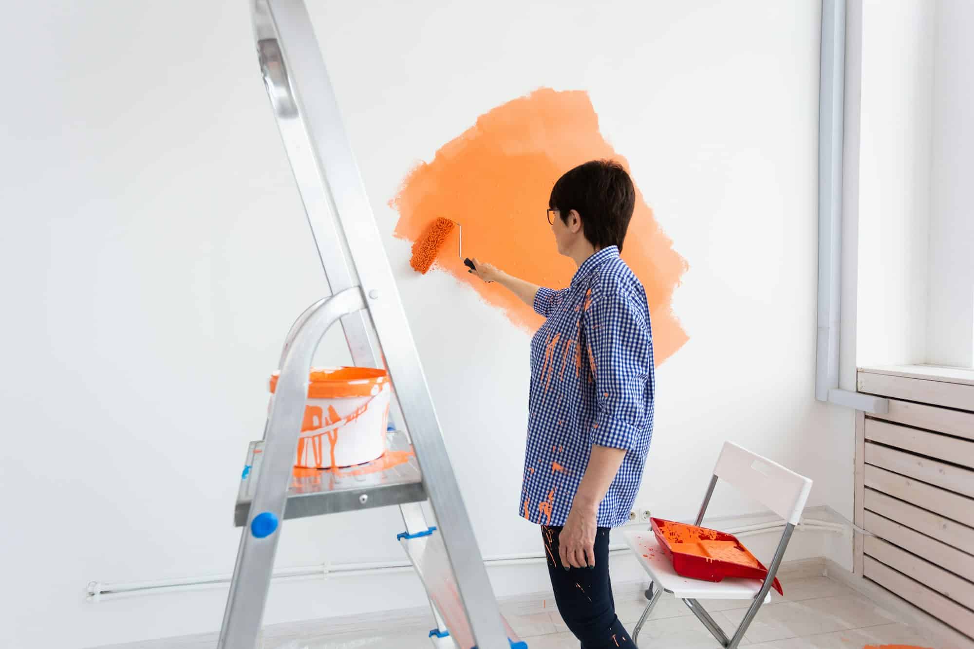 Pretty smiling middle-aged woman painting interior wall of home with paint roller. Redecoration